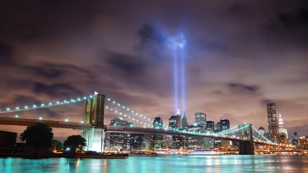 NYCOSH - Brooklyn Bridge at night with Manhattan skyline in the background and two blue light beams shining upward into the sky.