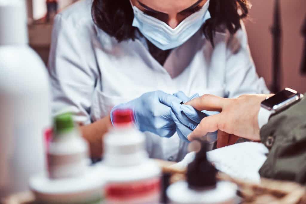 NYCOSH - A person wearing a mask and gloves gives a manicure to another person, with bottles of nail products visible in the foreground.
