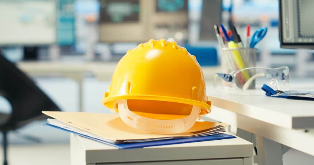 NYCOSH - A yellow hard hat resting on top of folders and documents on an office desk, with pens and safety goggles nearby.