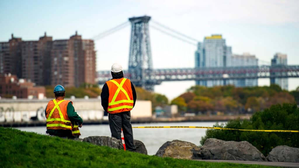 NYCOSH - Two construction workers wearing safety vests and helmets observe a riverfront area with a bridge and city buildings in the background.