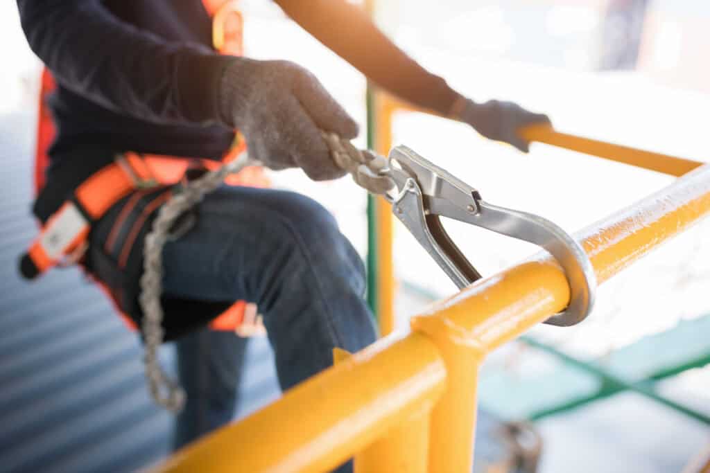 NYCOSH - Worker wearing safety harness and gloves attaches a lanyard hook to a yellow metal railing at a construction or industrial site.