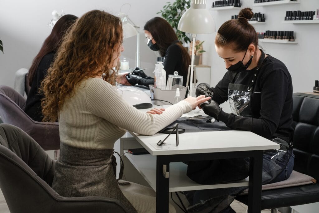 NYCOSH - A nail technician wearing gloves and a mask works on a customer's nails at a salon, with other clients and technicians visible in the background.