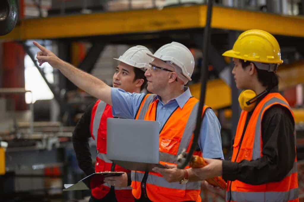 NYCOSH - Three workers in safety vests and helmets stand in an industrial setting; one points ahead, another holds a laptop, and the third holds equipment.