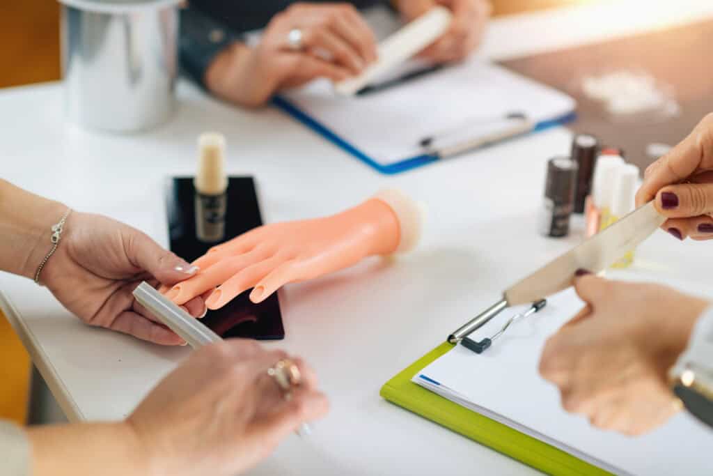 NYCOSH - Three people practice nail care techniques on a mannequin hand at a table with nail files, nail polish, and clipboards.