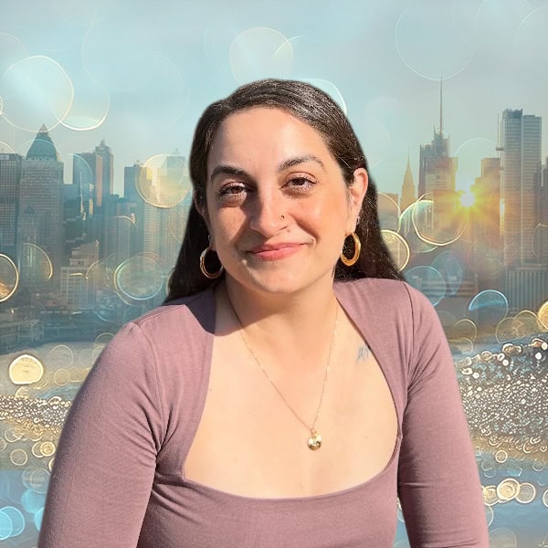 NYCOSH - A woman with long brown hair, wearing a mauve top and gold hoop earrings, sits in front of a city skyline with sunlight and light bokeh effects in the background.