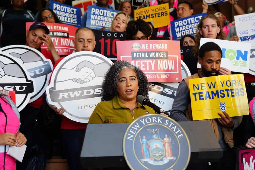 NYCOSH - A woman speaks at a podium with the New York State seal, surrounded by people holding signs supporting warehouse and retail worker safety, unions, and protections against wage theft.