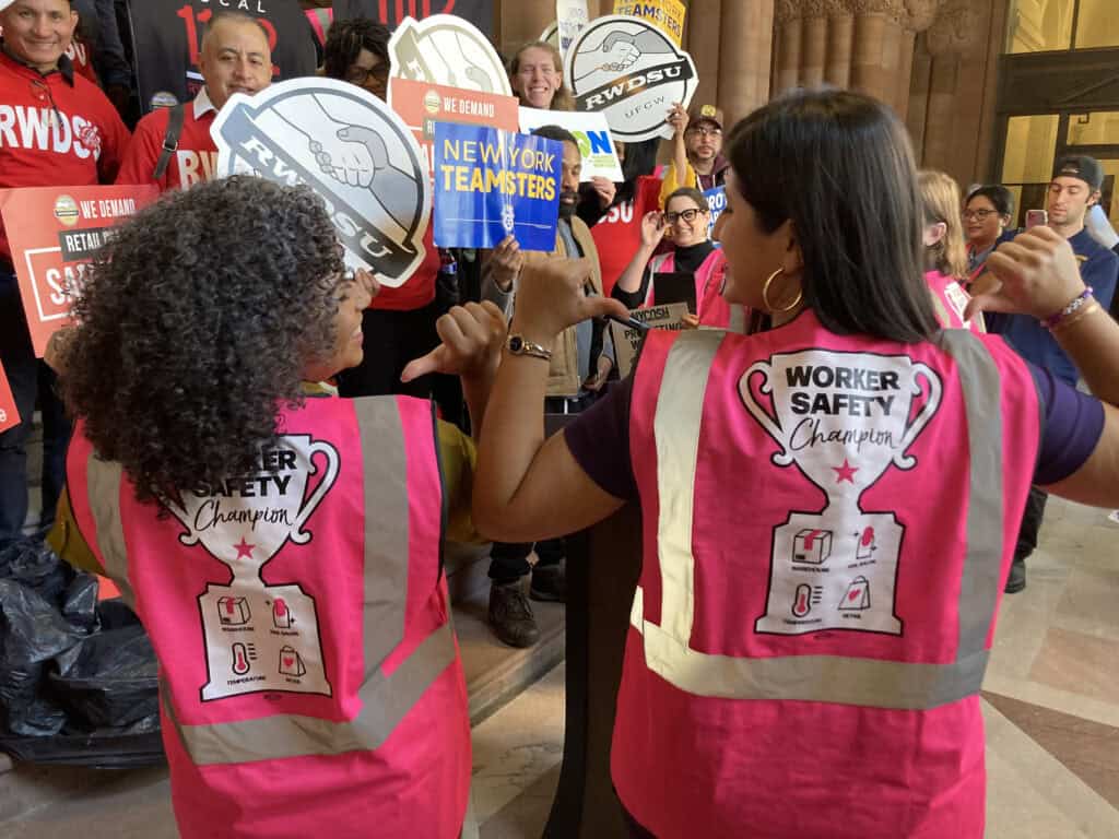 NYCOSH - Two people wearing bright pink "Worker Safety Champion" vests stand at a podium, raising their arms, while others hold union signs at a rally indoors—an inspiring moment about NYCOSH and worker safety advocacy.