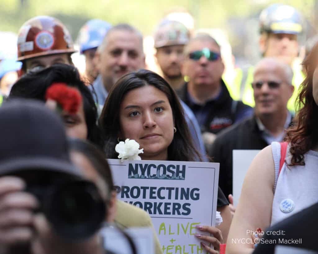NYCOSH - A woman holds a white flower and an NYOSH sign at a gathering; people wearing hard hats and onlookers stand in the background.