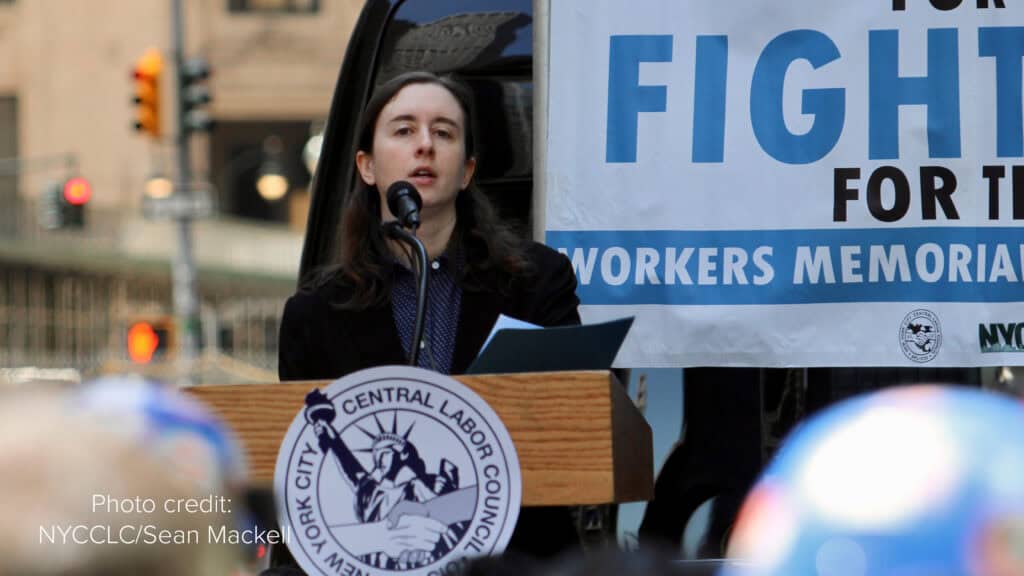 NYCOSH - A woman, an NYCOSH Member, speaks at a podium bearing the New York City Central Labor Council logo during a Workers Memorial event. A banner and a blurred crowd are visible in the background.