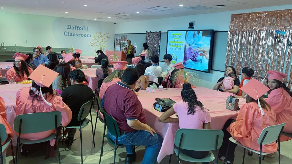 NYCOSH - People in pink graduation caps and gowns sit at tables in a decorated classroom labeled "Daffodil Classroom," watching a presentation on a screen at the front of the room.