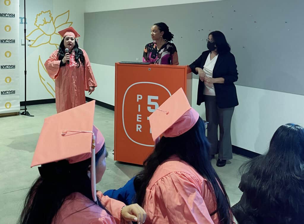 NYCOSH - A graduate in a pink cap and gown speaks at a podium while others, also in pink caps and gowns, listen. Two women stand behind the podium.