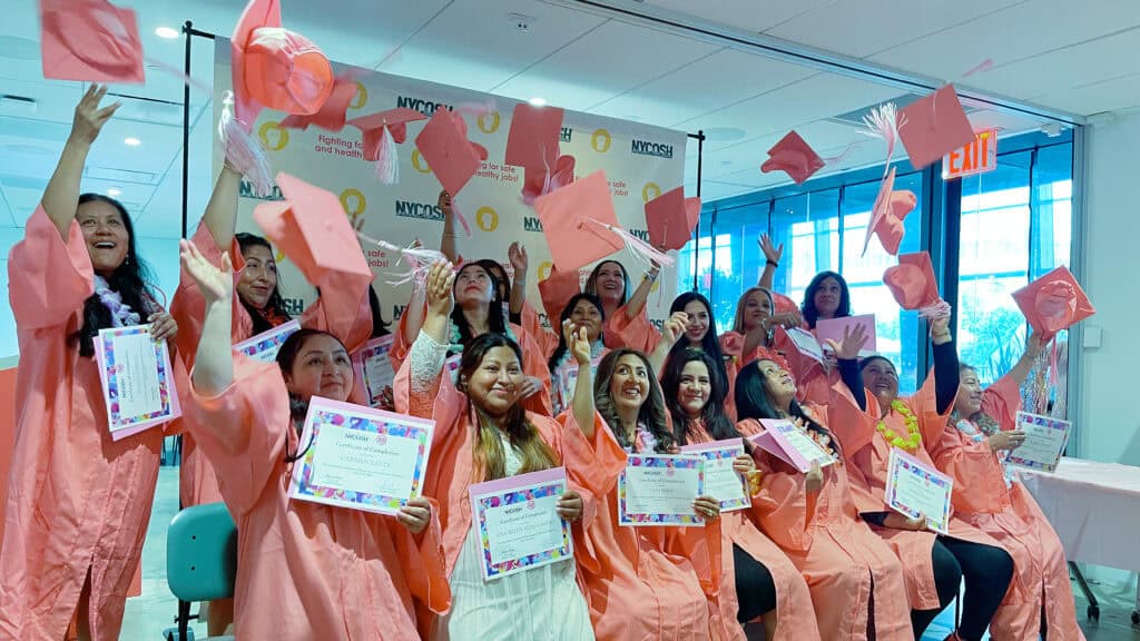 NYCOSH - A group of graduates in pink gowns celebrate by tossing their caps in the air while holding certificates in a brightly lit indoor setting.