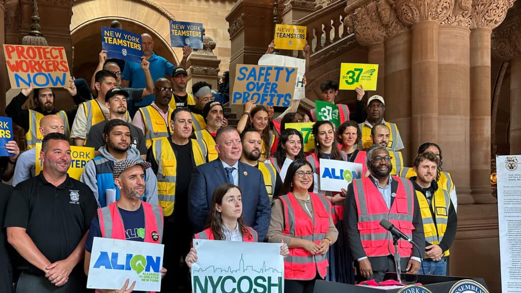 NYCOSH - A group of people in safety vests stand on steps holding signs like "Protect Workers Now" and "Safety Over Profits" at an indoor rally about NYCOSH, advocating for workplace health and safety.