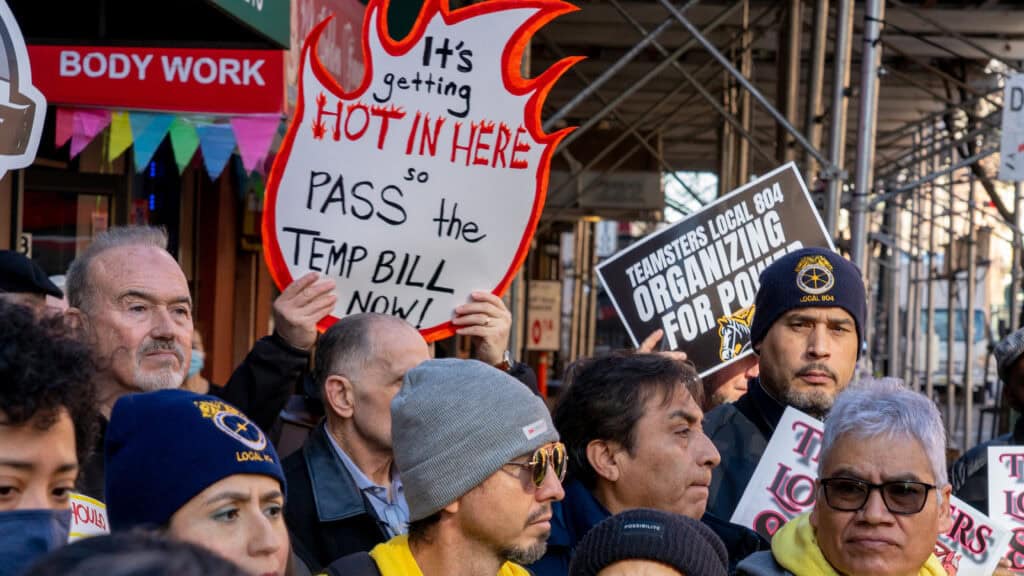 NYCOSH - A group of people with the Temp Coalition stand together at a protest, holding signs—including one reading "It's getting hot in here so PASS THE TEMP BILL NOW!"—and another supporting union organizing.