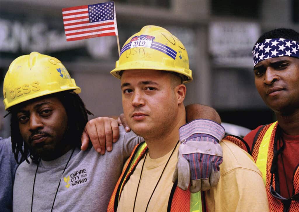 NYCOSH - Three construction workers in hard hats and safety vests stand together, one with an American flag on his helmet, looking at the camera—symbols of the resilience supported by programs like the WTC Health Program.