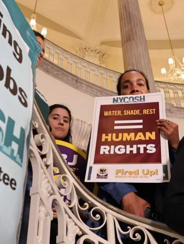 NYCOSH - Several people stand on a staircase holding signs advocating for workers' rights, including one that reads, "Water, Shade, Rest = Human Rights.