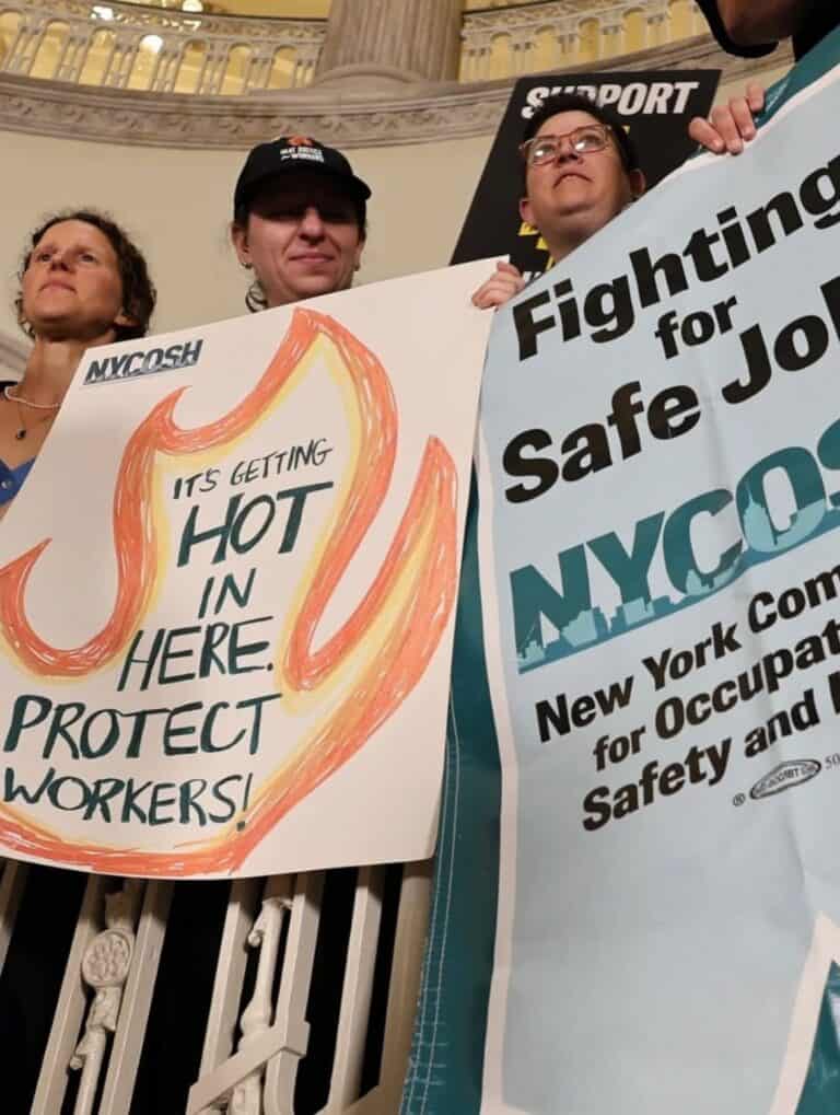 NYCOSH - Three people standing indoors hold signs advocating for worker safety; one sign reads "It's getting hot in here, protect workers!" and another promotes fighting for safe jobs.