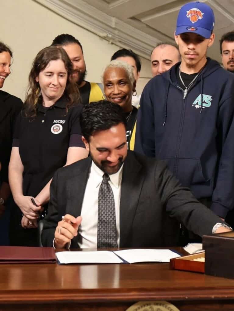 NYCOSH - A man in a suit sits at a desk signing documents while a group of people stand behind him, some smiling and watching the event.