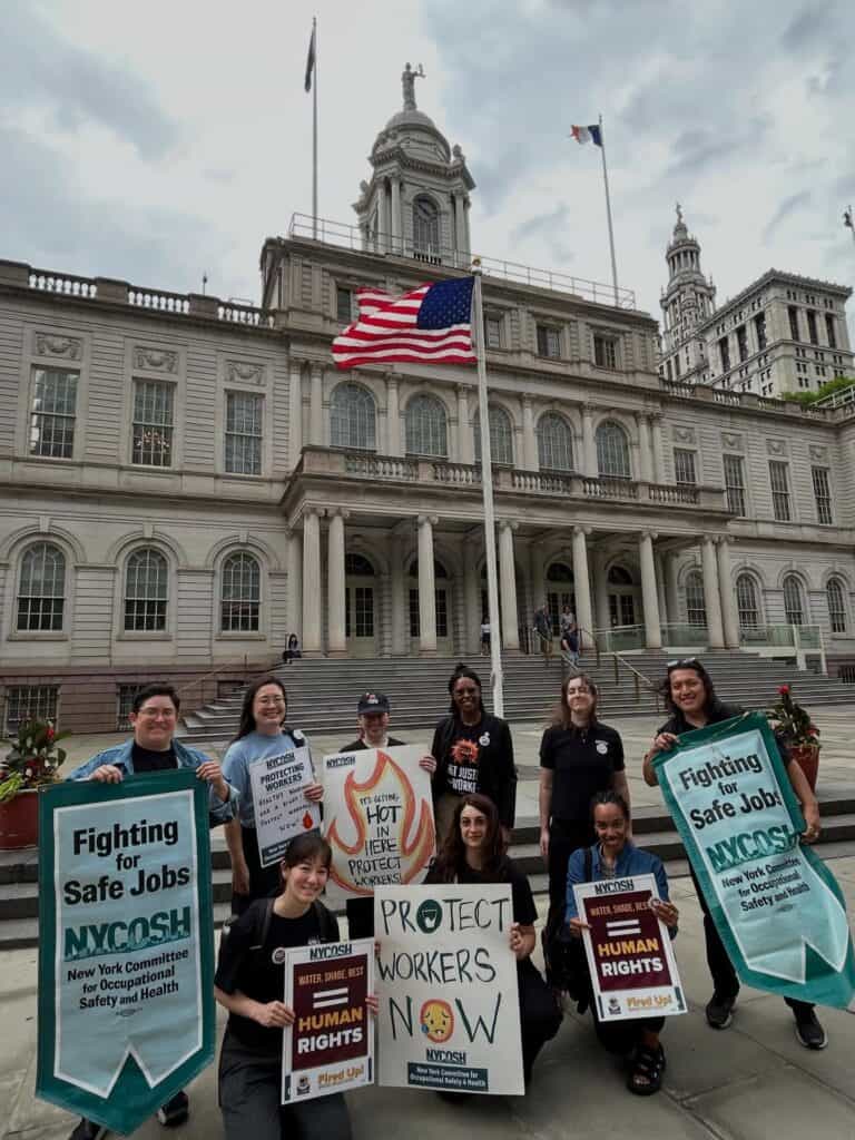 NYCOSH - A group of people stand in front of a government building holding signs that advocate for safe jobs, worker protection, and human rights. An American flag is visible above the entrance.