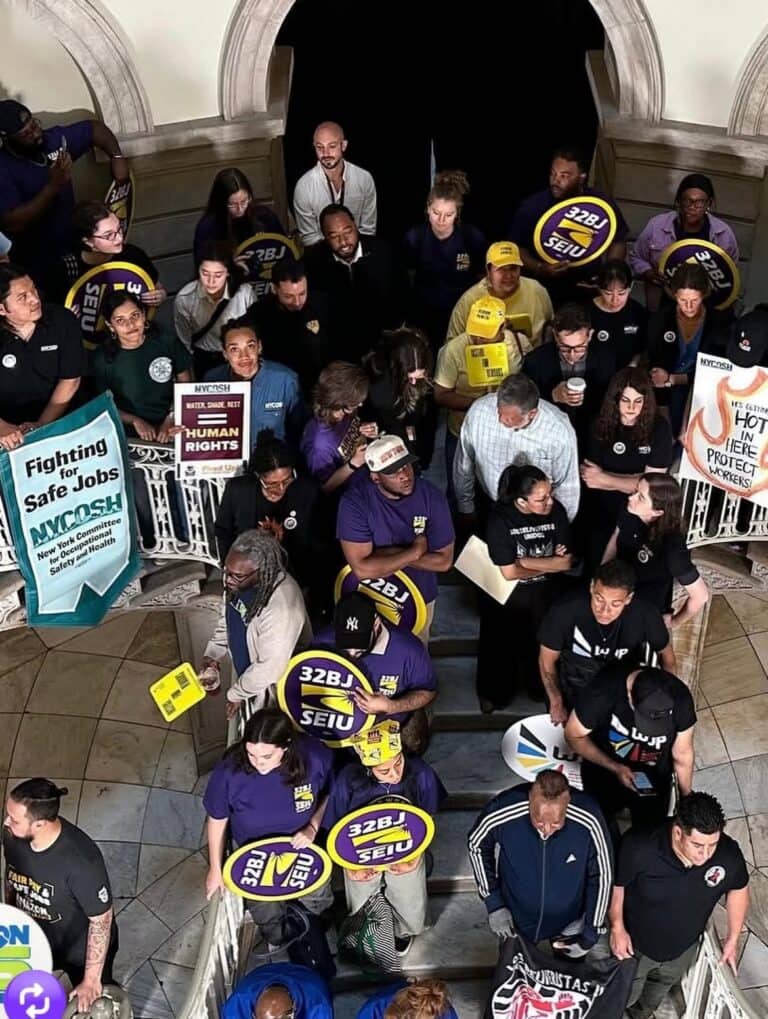 NYCOSH - A group of people stand closely together on stairs holding union signs and posters advocating for safe jobs and human rights.